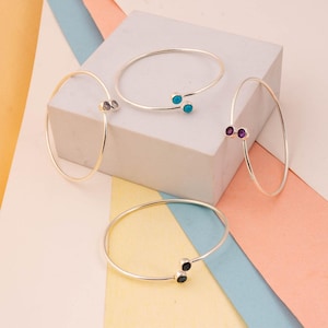 May include: Four delicate bangles with gemstone accents. The bracelets are silver-toned, with small, colourful stones in purple, teal, grey, and black. The bangles are displayed on a white and pastel-coloured surface.