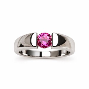 May include: A silver-toned metal ring featuring a single, round, vibrant pink gemstone set in a tension-style setting. The band is smooth and polished, with a solid, wide profile that tapers slightly towards the centre where the stone is held.