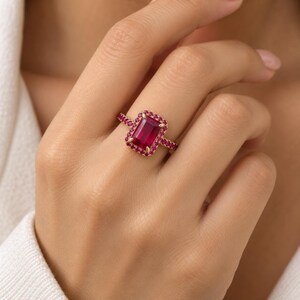 Ruby statement ring with emerald cut pink ruby gemstone, matching halo, and 925 sterling silver gold plated band worn on a model’s finger.