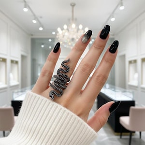 Puede incluir: Un anillo de serpiente negro con un diseño detallado. El anillo se envuelve alrededor del dedo, con un cuerpo texturizado y una cabeza de serpiente. El anillo se lleva en una mano con esmalte de uñas negro.