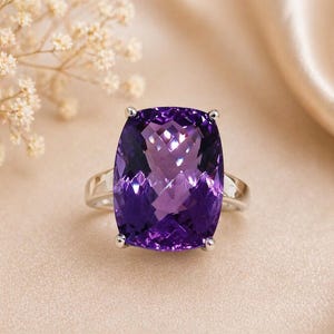 Pode incluir: Anel de prata com uma grande ametista de corte retangular. A pedra roxa profunda é fixada por quatro garras. O anel é apresentado sobre um fundo neutro, com iluminação suave, destacando a joia.