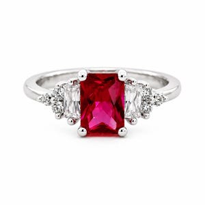 Puede incluir: Un anillo de tono plateado con una gran piedra central rectangular de color rojo vibrante. La piedra central está flanqueada por dos piedras transparentes de corte baguette rectangular y piedras de acento redondas y claras a los lados.