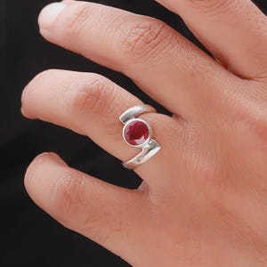 Puede incluir: Un anillo de plata con una gema roja grande y redonda. El anillo tiene un diseño moderno y asimétrico con una banda curva. La gema roja es el punto focal, engastada en un bisel simple.