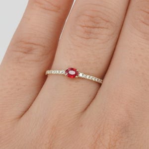 Op de afbeelding: Een gouden ring met een levendige rode ovale edelsteen in het midden, geflankeerd door kleinere, sprankelende witte stenen. De band is ook versierd met kleine witte stenen, wat een delicaat en elegant ontwerp creëert. De ring wordt op een vinger getoond.