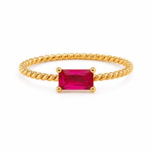 May include: A gold ring featuring a twisted rope-style band and a single rectangular baguette-cut vibrant pink gemstone set horizontally in the center with four small prongs holding the stone securely in place against the metallic gold finish.