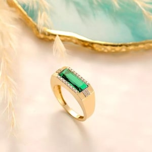 Può includere: Un anello d'oro con una pietra preziosa rettangolare verde smeraldo. L'anello è impreziosito da piccole pietre preziose trasparenti attorno allo smeraldo e sulle spalle. Sfondo sfocato.