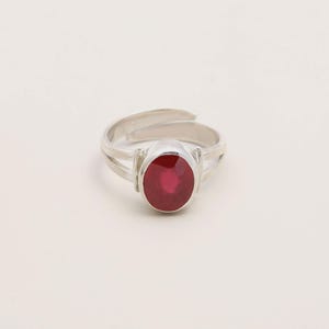 Op de afbeelding: Een zilveren ring met een ovale, dieprode edelsteen. De ring heeft een gespleten schachtontwerp en een verstelbare band. De edelsteen is het middelpunt, gezet in een eenvoudige, elegante setting. De ring wordt gepresenteerd tegen een effen witte achtergrond.