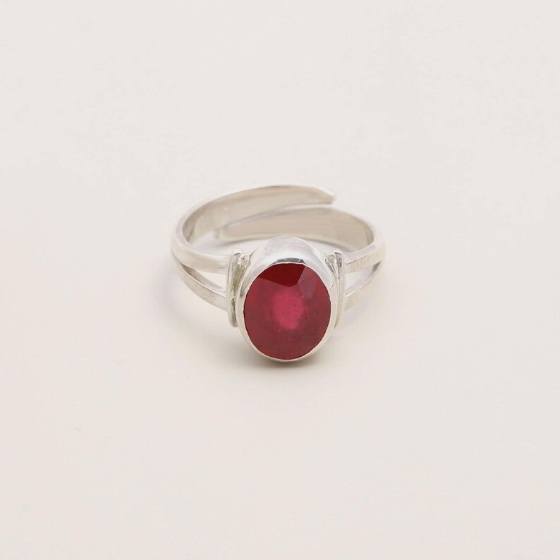 Op de afbeelding: Een zilveren ring met een ovale, dieprode edelsteen. De ring heeft een gespleten schachtontwerp en een verstelbare band. De edelsteen is het middelpunt, gezet in een eenvoudige, elegante setting. De ring wordt gepresenteerd tegen een effen witte achtergrond.