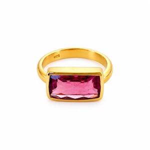 Può includere: Un anello color oro con una grande pietra preziosa magenta rettangolare sfaccettata, incastonata in una montatura in metallo liscio. L'interno dell'anello reca chiaramente il marchio 925, che indica la composizione metallica del gioiello.