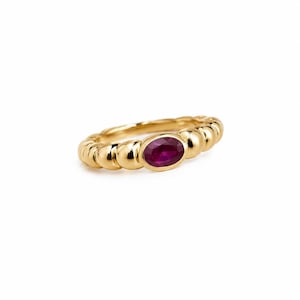 Op de afbeelding: Een gouden ring met een textuur van kralen en een enkele ovaal geslepen, diep roze-rode edelsteen in het midden. Het metaal heeft een gepolijste, reflecterende afwerking en de steen is stevig geplaatst in een gladde, afgeronde zetting.
