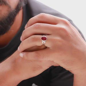 Op de afbeelding: Een zilveren ring met een ovale, dieprode edelsteen. De ring heeft een eenvoudig, elegant ontwerp dat de levendige kleur van de steen benadrukt. De ring wordt aan een vinger gedragen.