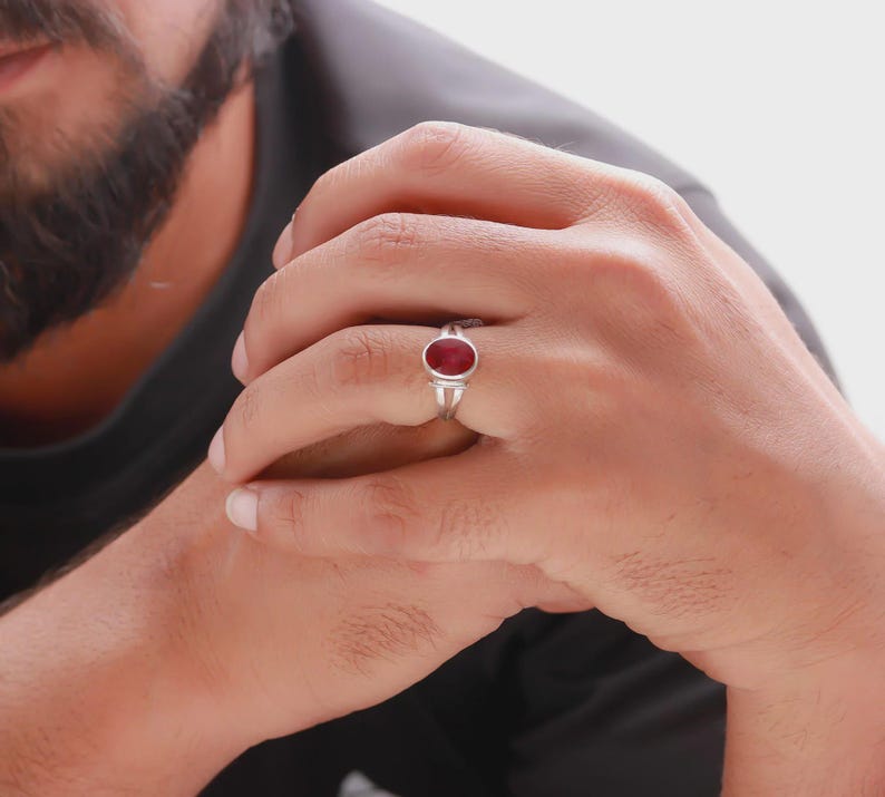Op de afbeelding: Een zilveren ring met een ovale, dieprode edelsteen. De ring heeft een eenvoudig, elegant ontwerp dat de levendige kleur van de steen benadrukt. De ring wordt aan een vinger gedragen.