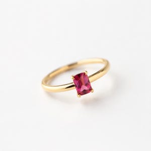 May include: A gold ring featuring a single rectangular emerald-cut pink gemstone set in a four-prong setting. The band is thin, smooth, and polished, showcasing a minimalist design that highlights the vibrant color of the central stone.