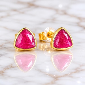 Può includere: Un paio di orecchini a bottone dorati con gemme rosa triangolari vibranti. Le gemme sono incastonate in una lunetta dorata. Gli orecchini sono esposti su una superficie riflettente.