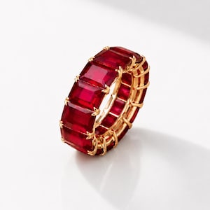 Può includere: Un anello a fascia eternity con una fila continua di pietre preziose rettangolari rosso scuro, taglio smeraldo, incastonate in una montatura in oro giallo lucido. Il design presenta una serie di pietre rosse vivaci fissate da piccole griffe.