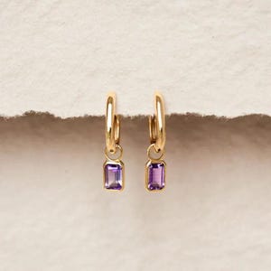 Könnte beinhalten: Ein Paar goldene Creolen, die jeweils mit einem kleinen, rechteckigen, smaragdgeschliffenen violetten Edelsteinanhänger versehen sind, der an der Unterseite des Reifens hängt. Die Ohrringe bestehen aus poliertem gelbem Metall.