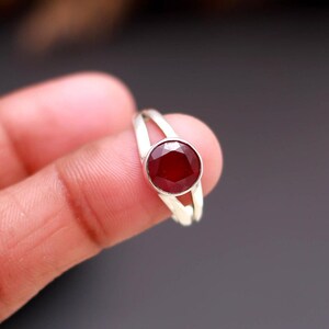 Op de afbeelding: Een zilveren ring met een ronde, gefacetteerde, dieprode edelsteen. De ring heeft een gespleten schachtontwerp, met twee parallelle banden die om de vinger buigen. De edelsteen is gezet in een bezel-zetting.