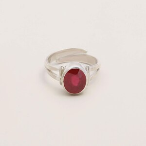 Op de afbeelding: Een zilveren ring met een ovale, dieprode edelsteen. De ring heeft een gespleten schachtontwerp en een verstelbare band. De edelsteen is het middelpunt, gezet in een eenvoudige, elegante setting. De ring wordt gepresenteerd tegen een effen witte achtergrond.