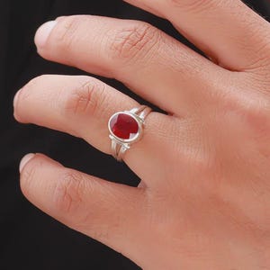 Op de afbeelding: Een zilveren ring met een grote, ovale, dieprode edelsteen. De ring heeft een gespleten schachtontwerp, waarbij twee banden samenkomen om de steen vast te houden. De edelsteen is het middelpunt, gezet in een eenvoudige, elegante stijl.