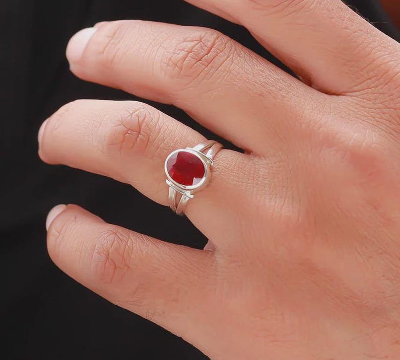 Op de afbeelding: Een zilveren ring met een grote, ovale, dieprode edelsteen. De ring heeft een gespleten schachtontwerp, waarbij twee banden samenkomen om de steen vast te houden. De edelsteen is het middelpunt, gezet in een eenvoudige, elegante stijl.