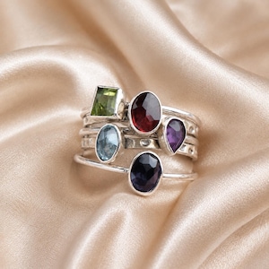 Pode incluir: Um conjunto de anéis de prata empilháveis com cinco pedras preciosas distintas de várias formas e cores, incluindo uma pedra verde quadrada, uma grande pedra vermelha oval, uma pequena pedra azul-clara oval, uma pedra roxa em forma de pera e uma pedra oval escura.