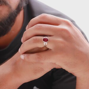 Op de afbeelding: Een zilveren ring met een ovale, dieprode edelsteen. De ring is gezet op een zilveren band en wordt aan een vinger gedragen. De achtergrond is neutraal.