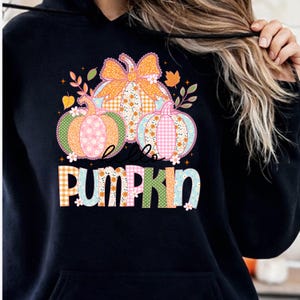 Puede incluir: Sudadera con capucha negra con un diseño de "Hello Pumpkin". El diseño incluye tres calabazas con patrones florales y a cuadros, y un gran lazo naranja. El texto es de varios colores y patrones.