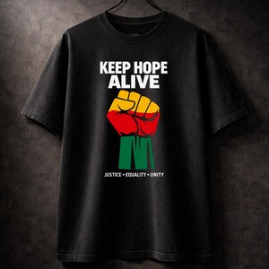 Può includere: T-shirt nera con la scritta "KEEP HOPE ALIVE" sopra un pugno chiuso rosso, giallo e verde. Le parole "JUSTICE • EQUALITY • UNITY" sono stampate sotto. La maglietta è appesa a una gruccia nera.