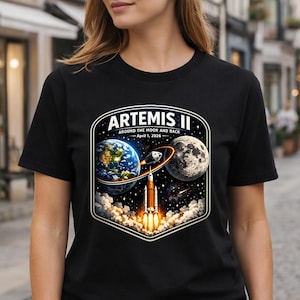 May include: Black t-shirt featuring a colorful graphic of a rocket launching from Earth towards the moon, with the text "ARTEMIS II" and "AROUND THE MOON AND BACK - April 1, 2026". The design includes Earth, the moon, and a rocket.