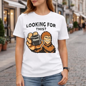 Könnte beinhalten: Weißes T-Shirt mit dem Text "LOOKING FOR THIS?" über einer Cartoon-Illustration eines lächelnden Bigfoot, der eine 10-mm-Stecknuss hält. Der Bigfoot ist braun und die Stecknuss ist schwarz.