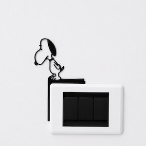May include: A black metal wall decal featuring the cartoon character Snoopy standing on the edge of a rectangular frame. The decal is positioned above a white light switch plate that contains three vertical black rectangular toggle switches.