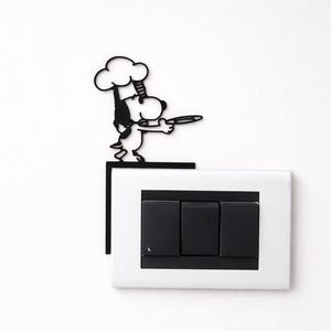 Könnte beinhalten: Schwarze Metall-Wanddekoration von Snoopy als Koch, der einen Teller hält, positioniert über einem weißen Lichtschalter mit drei schwarzen Schaltern. Der Koch trägt eine Kochmütze und eine Schürze. Die Wand ist weiß.