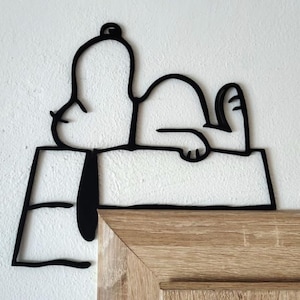 May include: Black metal Snoopy wall art. The artwork depicts Snoopy lying on top of his doghouse. The doghouse is a simple rectangular shape. The artwork is mounted on a white wall and a wooden surface.