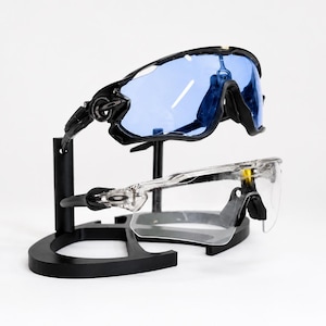 Puede incluir: Dos pares de gafas de sol deportivas exhibidas en un soporte negro. El par superior tiene montura negra y lentes azules, mientras que el par inferior tiene montura transparente. El soporte es negro y tiene una base curva.