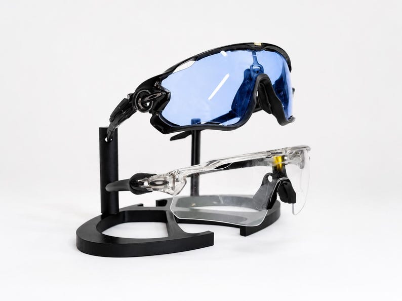 Puede incluir: Dos pares de gafas de sol deportivas exhibidas en un soporte negro. El par superior tiene montura negra y lentes azules, mientras que el par inferior tiene montura transparente. El soporte es negro y tiene una base curva.