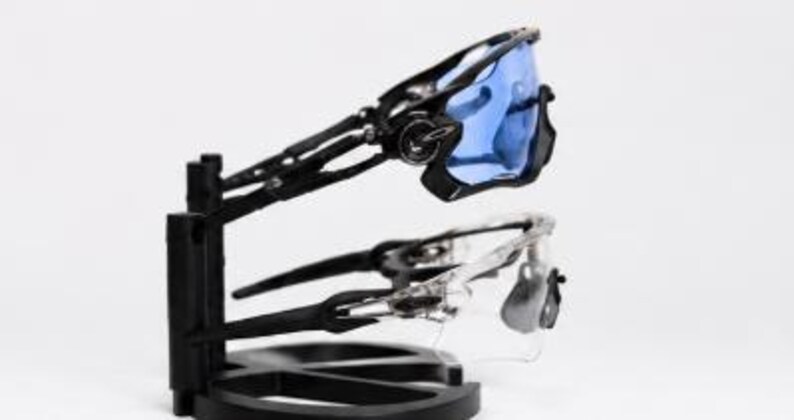 Puede incluir: Dos pares de gafas de sol deportivas expuestas en un soporte negro. Un par tiene montura negra y lentes azules, el otro montura transparente y lentes grises. El soporte tiene una base circular y un soporte vertical.