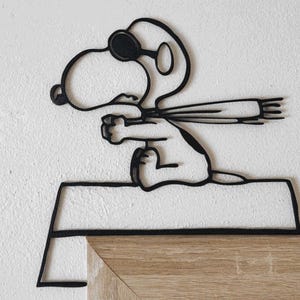 May include: Black metal Snoopy wall art. The artwork depicts Snoopy wearing goggles and a scarf, in a flying pose. The art is mounted on a white wall and a wooden surface.