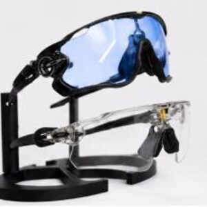 Puede incluir: Dos pares de gafas de sol deportivas expuestas en un soporte negro. Un par tiene montura negra con lentes azules, el otro montura transparente. Las gafas est&aacute;n dise&ntilde;adas para deportes y ofrecen protecci&oacute;n ocular.
