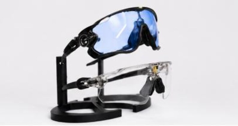 Puede incluir: Dos pares de gafas de sol deportivas expuestas en un soporte negro. Un par tiene montura negra con lentes azules, el otro montura transparente. Las gafas est&aacute;n dise&ntilde;adas para deportes y ofrecen protecci&oacute;n ocular.