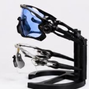 Puede incluir: Dos pares de gafas de sol deportivas se exhiben en un soporte negro. Un par tiene lentes azules y montura negra, el otro tiene lentes transparentes y montura plateada. El soporte es negro y tiene un dise&ntilde;o moderno.