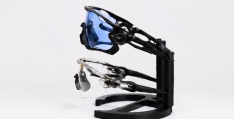 Puede incluir: Dos pares de gafas de sol deportivas se exhiben en un soporte negro. Un par tiene lentes azules y montura negra, el otro tiene lentes transparentes y montura plateada. El soporte es negro y tiene un dise&ntilde;o moderno.