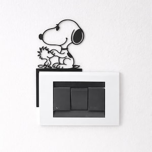 May include: Black metal Snoopy and Woodstock wall art above a white light switch plate with three black switches. The Snoopy figure is holding Woodstock. The wall is white.