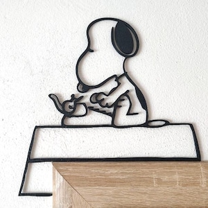 Può includere: Silhouette nera in metallo di Snoopy, seduto e con in mano una tazza da tè, appoggiata su una sporgenza rettangolare. L'opera d'arte è contro un muro bianco e una cornice in legno. Il design è una rappresentazione minimalista in stile cartone animato.