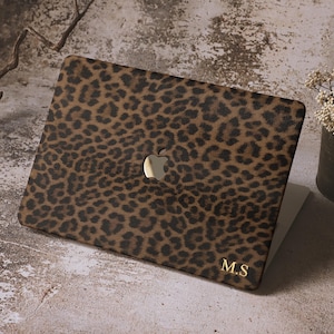 Könnte beinhalten: Eine Laptop-Hülle mit Leopardenmuster in Braun- und Schwarztönen. Die Hülle hat ein goldenes Apple-Logo und goldene Initialen "M.S". Der Laptop ist geöffnet und zeigt einen weißen Bildschirm.