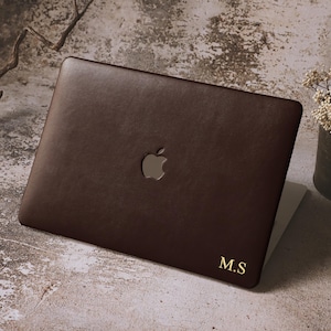 May include: A dark brown laptop case with the Apple logo cutout. The case has a smooth texture and gold-colored initials "M.S" in the bottom right corner. The laptop case is on a textured surface with a small vase of dried flowers in the background.