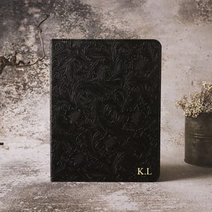 May include: A black rectangular notebook cover featuring an embossed floral pattern across the entire surface. The bottom right corner displays the gold-coloured initials K.L. The cover has a smooth texture and a consistent dark black finish.