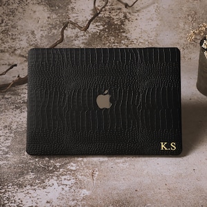Pode incluir: Capa preta para laptop com um padrão texturizado semelhante à pele de crocodilo. O logotipo da Apple é visível, juntamente com as iniciais douradas "K.S". A capa está sobre uma superfície de tons neutros.
