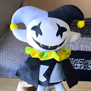 May include: A plush doll of a character with a white face, black star-shaped eyes, and a wide smile. The doll has a blue and black jester hat with yellow pom-poms, a green and yellow ruffled collar, and a black cape. The doll's body is white with a gray and black emblem.