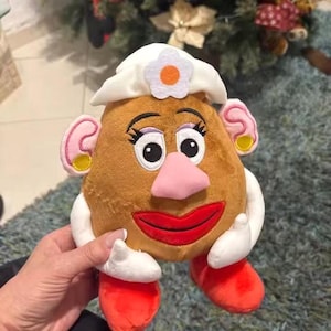 May include: A plush toy resembling Mrs. Potato Head, featuring a brown potato-shaped body with a white hat, pink nose, and red lips. The toy has large black eyes, pink ears with yellow earrings, and red feet. The arms are white with small white hands.