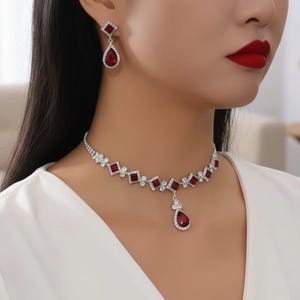 Può includere: Un set di collana e orecchini argentati con dettagli di pietre preziose rosse. La collana presenta un design geometrico con pietre quadrate e a goccia. Gli orecchini riprendono il design della collana.